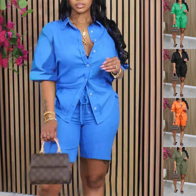 Stylish women wearing colorful short sleeve shirt and shorts set with drawstring waist, accessorized with gold jewelry, holding a designer handbag, standing against wooden panel background.
